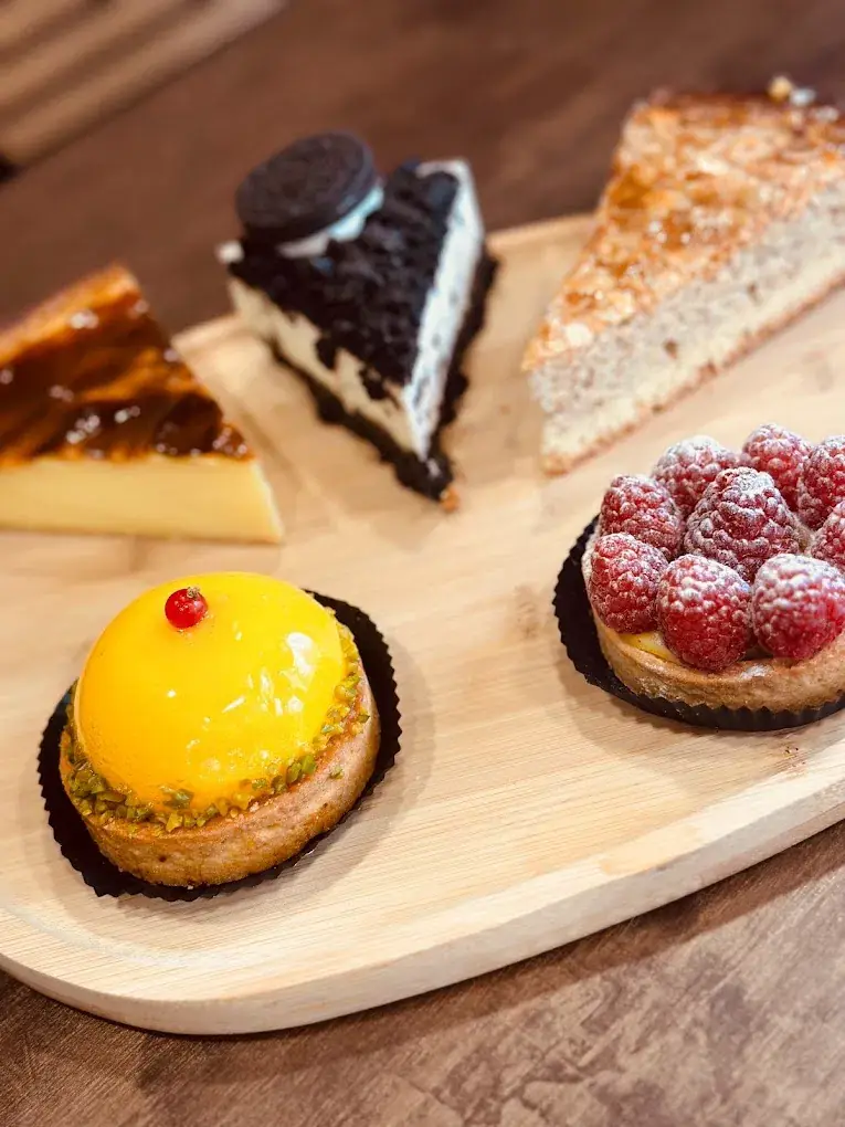 Quatre pâtisseries sur une planche en bois : une tarte aux framboises, un dôme à la mangue, une part de cheesecake et un gâteau caramélisé
